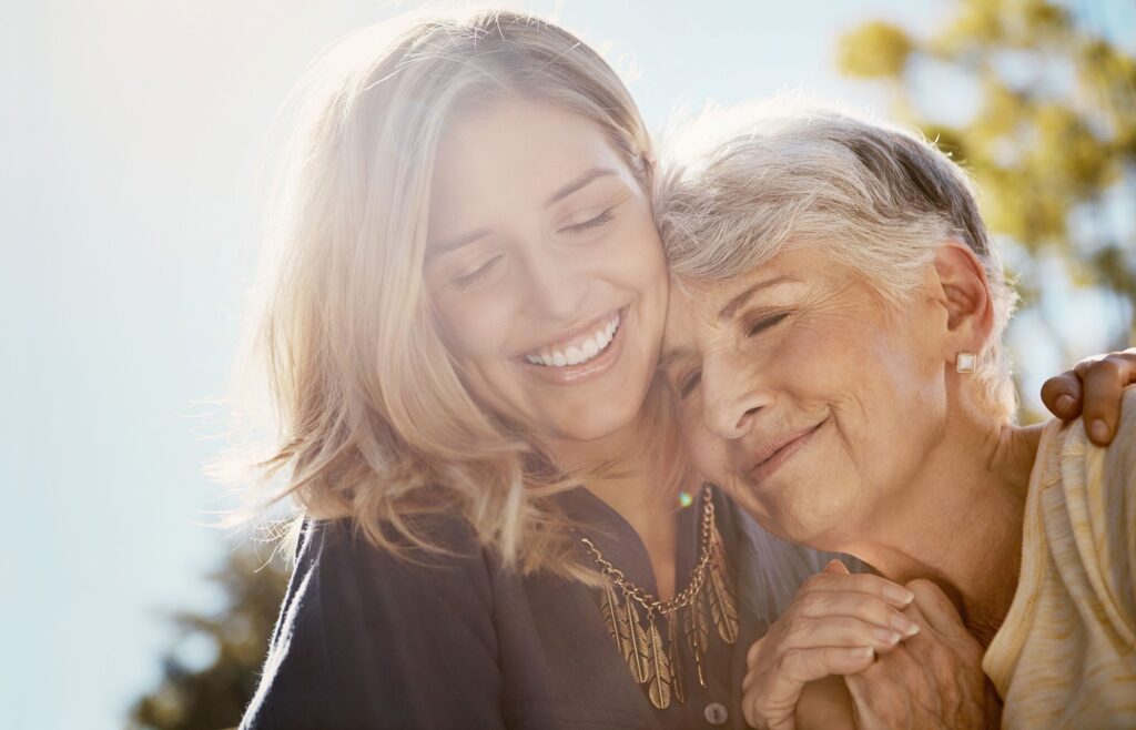 Older woman with short gray hair leaning on and hugging younger woman in the sunshine
